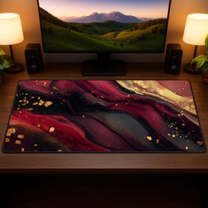 Può includere: Tappetino da scrivania rettangolare con un design astratto nei toni del rosso, nero e oro. Il tappetino presenta un effetto marmorizzato con spruzzi dorati. È posizionato su una scrivania in legno, con un monitor del computer e altoparlanti sullo sfondo.