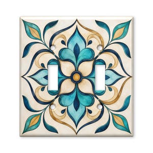 May include: A square double light switch wall plate featuring a symmetrical floral pattern in shades of teal, dark blue, and beige. The design centers on a circular gold accent, with two vertical rectangular cutouts for switches and four screw holes.