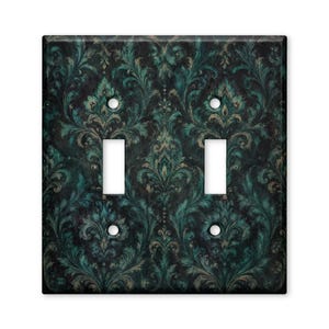 May include: A double light switch wall plate featuring a dark black background with a repeating teal and muted gold damask pattern. The rectangular cover includes two vertical rectangular cutouts for switches and four circular screw holes.