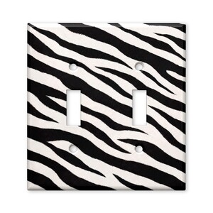 May include: A square double light switch wall plate featuring a black and white zebra stripe pattern. The cover includes two rectangular cutouts for toggle switches and four circular holes for mounting screws, arranged in a standard configuration.