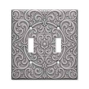 May include: A double light switch wall plate featuring a raised, silver-toned metallic finish with an intricate, symmetrical scrollwork pattern. The design includes a central floral motif surrounded by swirling filigree and four screw holes.