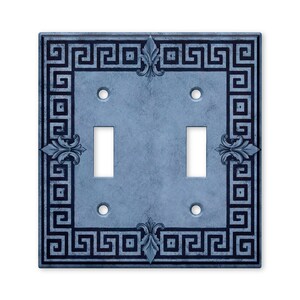 May include: A double light switch wall plate featuring a light blue, mottled stone-like texture. The border is decorated with a dark blue Greek key pattern and four symmetrical fleur-de-lis accents centered on each side of the square frame.