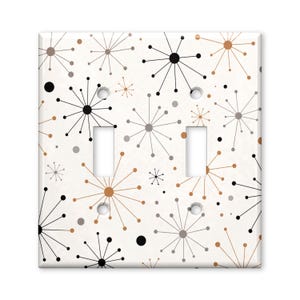 May include: A double light switch wall plate featuring a white background with a repeating pattern of black, gray, and copper-colored starburst shapes and small dots. The rectangular plate includes two vertical cutouts for toggle switches.