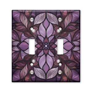 May include: A double light switch wall plate featuring a symmetrical floral mandala pattern in shades of purple, lavender, and deep plum. The design includes intricate leaf-like shapes and small white circular accents around the switch openings.