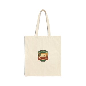 May include: A cream-coloured tote bag featuring a graphic of a living room scene within a national park-style emblem. The emblem reads "Living Room National Park" and "Certified Indoorist". The bag has two long handles.