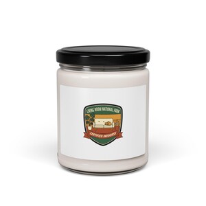 May include: A white candle in a glass jar with a black lid. The label features a green, brown, and red graphic with the text "LIVING ROOM NATIONAL PARK" and "CERTIFIED INDOORSY".