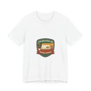 Könnte beinhalten: Weißes T-Shirt mit einem Aufdruck, der eine Wohnzimmerszene mit Sofa, Katze und Topfpflanze zeigt. Die Grafik befindet sich in einem Schild mit dem Text "LIVING ROOM NATIONAL PARK" und "CERTIFIED INDOORSY" auf einem roten Banner.
