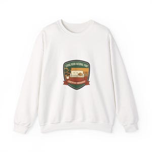 Könnte beinhalten: Weißes Sweatshirt mit Rundhalsausschnitt und einem grafischen Aufnäher. Der Aufnäher zeigt eine Cartoon-Wohnzimmerszene mit dem Text "LIVING ROOM NATIONAL PARK" und "CERTIFIED INDOORSY". Das Design umfasst ein Sofa, eine Katze und Topfpflanzen.