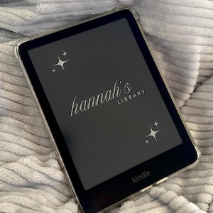 Puede incluir: Un lector de libros electrónicos Kindle negro con una funda protectora transparente. La pantalla muestra el texto "hannah's LIBRARY" en cursiva, con gráficos de estrellas. El logotipo de Kindle es visible en la parte inferior.