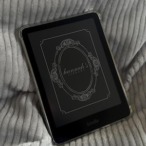Puede incluir: Un lector de libros electrónicos Kindle negro con una funda protectora transparente. La pantalla muestra el texto "hannah's LIBRARY" dentro de un marco adornado. La palabra "kindle" está impresa en la parte inferior del dispositivo.