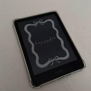 Puede incluir: Lector de libros electrónicos Kindle negro con una funda protectora transparente. La pantalla muestra un marco decorativo gris con lazos y el texto "hannah's KINDLE". El lector está sobre una superficie blanca.