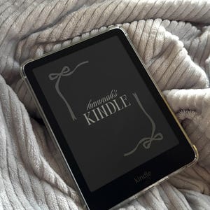 May include: A black Kindle e-reader in a clear case. The screen shows the text "hannah's KINDLE" in a decorative font, with bow and ribbon designs. The Kindle logo is at the bottom. The e-reader rests on a grey, textured surface.