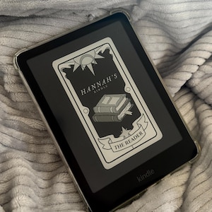 Puede incluir: Un lector de libros electrónicos Kindle negro con una funda protectora transparente. La pantalla muestra un gráfico con el texto "HANNAH'S KINDLE" y "THE READER" y una ilustración de libros. El lector está sobre una superficie gris texturizada.