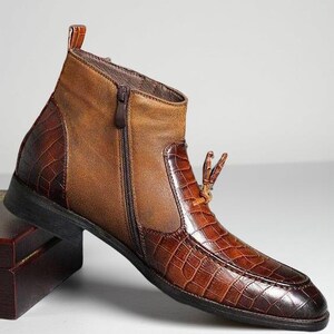 May include: A brown leather ankle boot with a side zipper and a textured, crocodile-like pattern on the toe and heel. The boot has a small pull tab at the top and a decorative leather detail. The boot is set on a black sole.