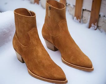 Puede incluir: Un par de botines de ante marrón con puntera y tacones de bloque de madera. Las botas tienen una cremallera lateral y están sobre un fondo nevado. Las botas son de un tono cálido y terroso.