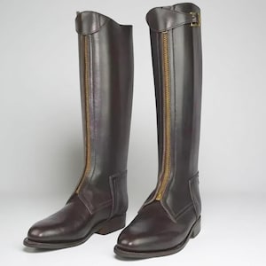 Könnte beinhalten: Ein Paar dunkelbraune Reitstiefel. Die Stiefel haben einen goldenen Reißverschluss vorne und eine Schnalle oben. Die Lederstiefel haben ein klassisches Design, ideal für den Reitsport. Die Stiefel sind etwa 40 cm hoch.