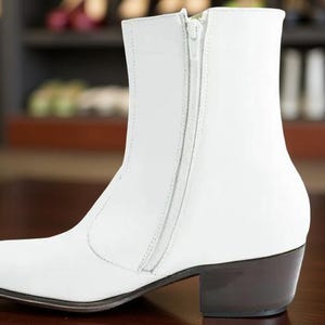 May include: A white ankle boot with a side zipper and a dark brown heel. The boot has a pointed toe and a smooth leather-like texture. The background is blurred, suggesting a store setting.