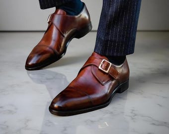 May include: Brown leather monk strap shoes with a polished finish. The shoes feature a single buckle closure and are worn with navy blue socks and pinstripe trousers. The shoes are a classic style.
