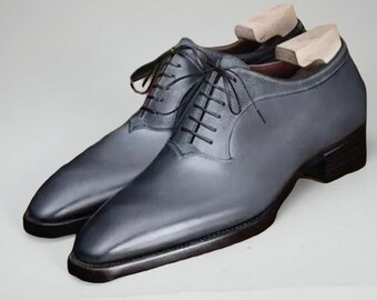 May include: A pair of gray leather dress shoes with black laces and wooden shoe trees. The shoes have a classic Oxford design with a polished finish and a dark sole. The footwear is displayed on a light gray background.