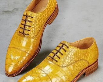 May include: A pair of yellow leather dress shoes with a textured, crocodile-like pattern. The shoes have brown laces and soles. The shoes are displayed on a light gray surface.