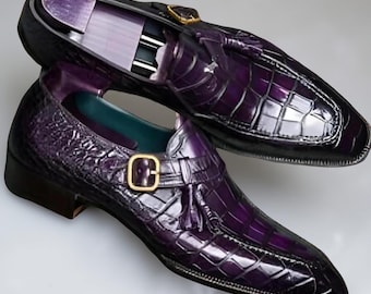 May include: A pair of purple leather dress shoes with a crocodile skin texture. The shoes feature a buckle and tassel detail, with a gold-coloured buckle. The shoes are displayed on a white surface.