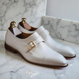 Może przedstawiać: Para białych sk&oacute;rzanych but&oacute;w z zapięciem typu monk strap ze złotymi klamrami. Buty mają zaokrąglony nosek i są prezentowane z drewnianymi prawidłowami. Podeszwy są brązowe, na tle marmuru.