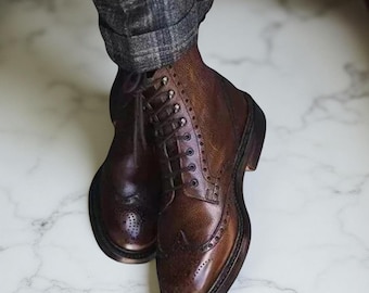 May include: A pair of brown leather brogue boots with a lace-up design. The boots feature a classic wingtip style with decorative perforations and a robust sole. The leather has a polished finish, and the boots are paired with grey plaid trousers.