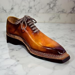 以下が含まれることがあります： 四角いトゥと装飾的なロープトリムが付いた、茶色の革製ドレスシューズ。靴はグラデーションカラーで、明るい茶色からつま先に向かって濃い色合いに変化します。靴紐は薄茶色とシルバーです。