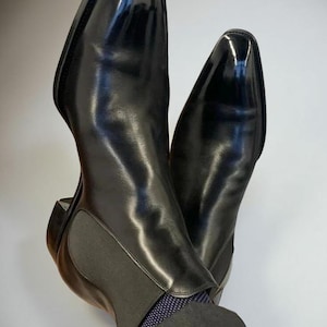 May include: A pair of black leather Chelsea boots with a high-gloss finish. The boots have a sleek, pointed toe and elastic side panels. The image shows the boots from a low angle, highlighting their polished appearance and design.