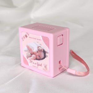 Peut inclure: Un appareil rose en forme de boîte avec une image de bébé et le texte "Welcome Baby" et "It's a Girl". Il possède une dragonne rose et un bouton. Le dessus comporte des fentes horizontales. Il s'agit probablement d'une étiqueteuse.