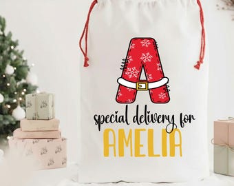 May include: White Christmas gift bag with red drawstring closure. The bag features a red letter "A" in a Santa-style design with snowflakes, a white belt, and the text "special delivery for AMELIA" in black and yellow.