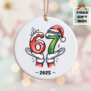 May include: A white ceramic Christmas ornament with a gold string. The ornament features the numbers 6 and 7 in red and green, held by cartoon hands. The text "THE YEAR OF" and "2025" are also present. The number 7 has a Santa hat with the name "BENJAMIN".