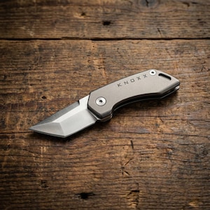 May include: A small, silver folding knife with a straight blade and the word "KNOXX" engraved on the handle. The knife is open, revealing the sharp blade. The knife is on a wooden surface.