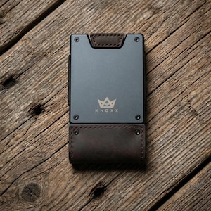 May include: A gray metal wallet with brown leather accents. The wallet has the word "KNOXX" and a crown logo printed on the front. The wallet is rectangular and has a bi-fold design.