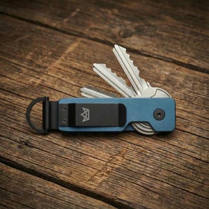 May include: A blue key organiser with a black clip and a key ring. The organiser holds several silver keys. The black clip has a logo. The key organiser is on a wooden surface.
