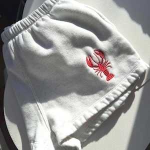 May include: Light gray sweat shorts with a red embroidered lobster design on the front. The shorts have an elastic waistband and a relaxed fit, perfect for casual wear. The shorts are made of a soft, comfortable material.