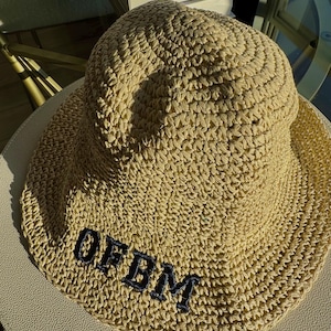 May include: A beige woven sun hat with a wide brim. The hat features the embroidered letters "OFBM" in navy blue on the front. The hat is made of a textured material and is sitting on a light-colored surface.