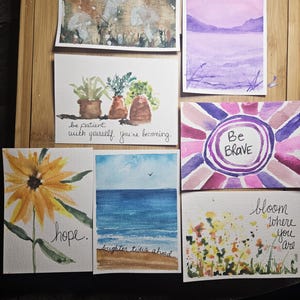 May include: A collection of seven watercolor paintings on paper. Designs include mushrooms, a purple landscape, potted plants with the text 'be patient with yourself. you're becoming.', a sunburst with 'Be Brave', a yellow flower with 'hope.', a beach scene with 'brighter tides ahead.', and flowers with 'bloom where you are'.