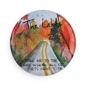 以下が含まれることがあります： 湖へと続く道を描いた水彩画が描かれた丸いボタン。オレンジと緑の紅葉が描かれています。「The Lakes」と「Take me to the lake where all the poets went to die」というテキストがボタンに書かれています。