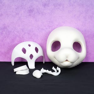 May include: A collection of white, unfinished fursuit parts, including a head, a helmet-like structure, and various other components. The head features large eye openings and a smiling expression. The items are arranged on a black surface against a purple backdrop.