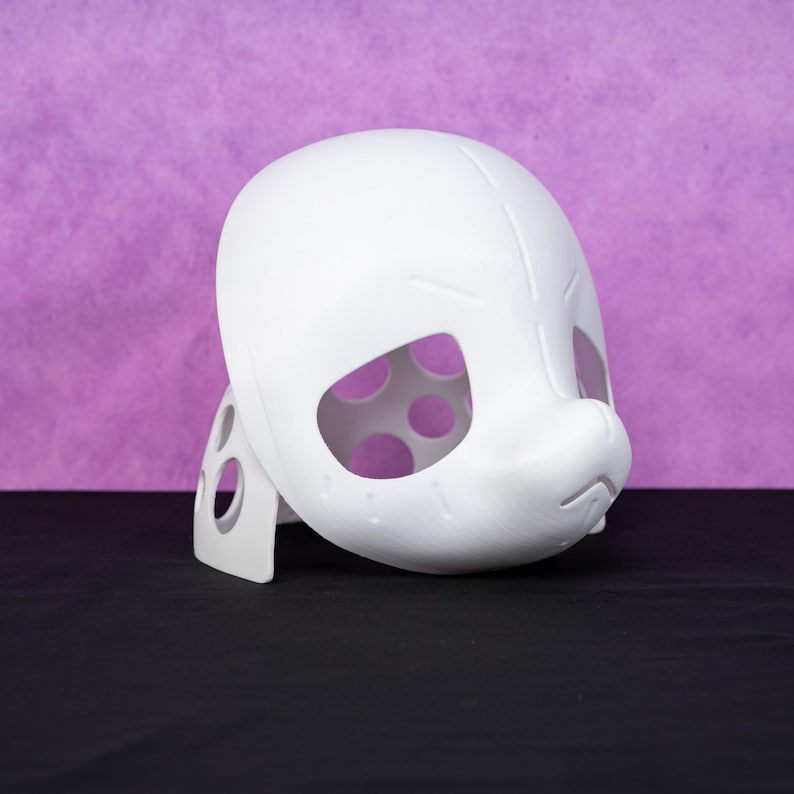 Puede incluir: Una m&aacute;scara blanca impresa en 3D con grandes aberturas para los ojos y una nariz prominente. La m&aacute;scara est&aacute; sostenida por un soporte blanco, y las aberturas de los ojos revelan un interior p&uacute;rpura con recortes circulares. El fondo es morado liso.