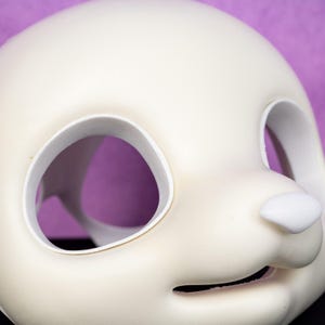 May include: A close-up of a white, blank mask with large eye openings and a small nose. The mask has a smooth surface and is likely made of plastic or a similar material. The background is a solid purple color.