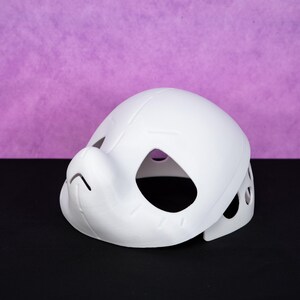 Puede incluir: Una m&aacute;scara blanca, impresa en 3D, con forma redondeada, con aberturas para los ojos y la boca. La m&aacute;scara tiene una nariz prominente y est&aacute; dise&ntilde;ada con varios agujeros peque&ntilde;os en los lados. El fondo es de color p&uacute;rpura s&oacute;lido.