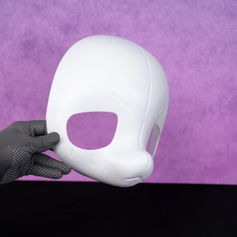 Puede incluir: Una m&aacute;scara blanca con grandes aberturas para los ojos y una nariz redondeada. La m&aacute;scara est&aacute; sujeta por una mano enguantada sobre un fondo morado. La m&aacute;scara parece estar hecha de un material liso y duro.