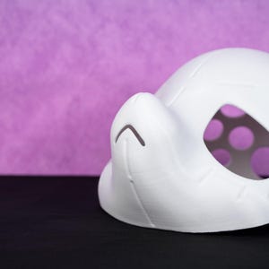 Puede incluir: Una m&aacute;scara blanca impresa en 3D con forma redondeada y una nariz prominente. La m&aacute;scara tiene una serie de peque&ntilde;os recortes circulares cerca del &aacute;rea de los ojos y un detalle de l&iacute;nea curva en la nariz. El fondo es morado liso.