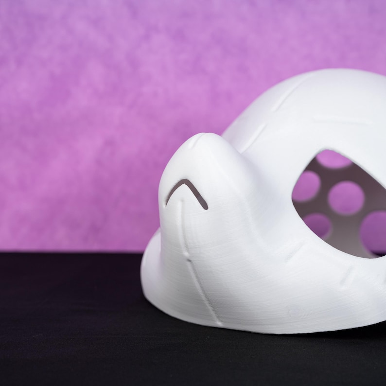 Puede incluir: Una m&aacute;scara blanca impresa en 3D con forma redondeada y una nariz prominente. La m&aacute;scara tiene una serie de peque&ntilde;os recortes circulares cerca del &aacute;rea de los ojos y un detalle de l&iacute;nea curva en la nariz. El fondo es morado liso.