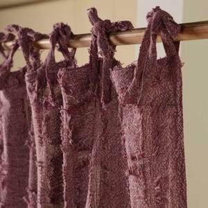 May include: Close-up of several dusty rose curtains hanging from a wooden rod. The curtains have a textured, woven appearance with knotted tie tops and frayed edges, creating a rustic aesthetic. The background is a neutral beige.