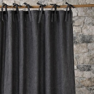 May include: A dark gray curtain hanging from a wooden rod with fabric ties. The curtain is made of a textured fabric and hangs in vertical folds. The background is a stone brick wall.