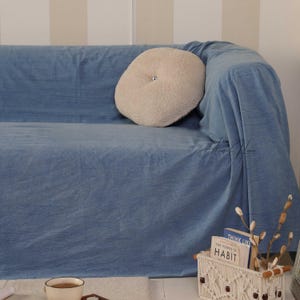 Może przedstawiać: Niebieska sofa pokryta niebieskim materiałowym narzutą. Na sofie znajduje się okrągła, beżowa, puszysta poduszka z guzikiem. Biały kosz z makramy zawiera książki i dekoracyjne gałęzie. Filiżanka kawy znajduje się na pierwszym planie.