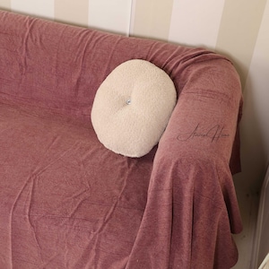 May include: A cream-coloured, round decorative cushion with a button centre rests on a mauve-coloured sofa. The sofa is covered with a matching mauve throw. The background features a striped wall.